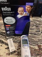 Photo showing Braun ThermoScan 7 open box packaging with image of mother and baby, and thermometer and box of filters displayed on a patterned surface.