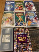 Top view of the 7 Disney VHS tapes and the Disney Who's Who book arranged on a surface, displaying the covers of each VHS sleeve and the book's front cover.