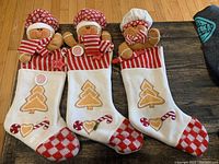 Front view of three gingerbread-themed Christmas stockings arranged on a wooden floor, showing white fabric bodies with red and white checkered toes and cuffs, embroidered gingerbread trees, peppermint candy, and plush gingerbread man tops with red plaid and white chef hats.