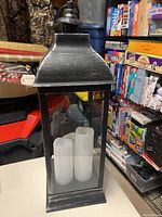 Full view of the large black metal and glass lantern with three white LED candles inside. Handle of lantern visible and appears damaged.