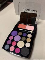 Close-up photo of one Samani Multi Nuances makeup palette opened showing 16 eyeshadows, 2 blushes in coral and pink, double-ended applicator brush, and mirror inside a black compact with transparent lid.