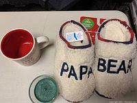 Photo showing white fluffy Papa Bear slippers, a cream mug with red interior, and a green evergreen scented candle in a glass jar.