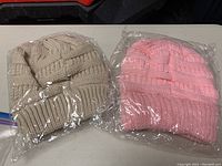 Two pony tail toques, pink and beige, sealed in plastic bags on a flat surface