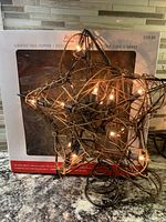 Rustic star-shaped tree topper lit with clear lights in front of its original box.