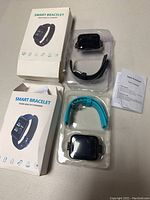 Two smart bracelets in plastic trays with their respective boxes and user manual. Black and teal wristbands are visible.