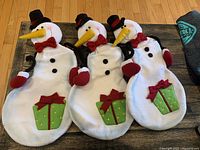 Three white snowman-shaped Christmas stockings lying side by side on a wooden floor, each with red mittens, black top hats, red bow ties, black buttons, and green gift box appliques with red bows and yellow dots.