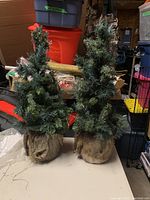 Full view of two artificial pine trees side by side showing green needles and burlap bases on a cluttered background.