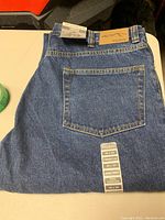Back view of folded blue denim jeans showing a single back pocket and attached size tags.