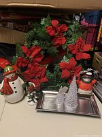Photo showing 18 inch homemade green wreath decorated with red poinsettia garland, two ceramic snowman figurines, cardinal statue, two clear Christmas tree figurines on a silver metal charger plate.