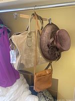 Beige leather duffel bag, brown velvet hat with feathers, and tan leather satchel hanging on hanger