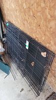 Black metal folding wire dog crate folded flat and leaning against a wooden wall, showing the front door and wire frame.