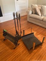 Photo showing fireplace accessory stand with poker, shovel, and tongs, next to the firewood holder on wood floor.