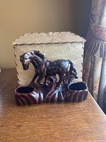 Front view of brown glazed ceramic horse lamp with ornate details and two hollow sections on the base, sitting in front of a beige textured background.