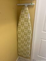 Full view of folding ironing board with fabric cover featuring palm tree checkered pattern hung on a wall.