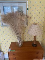 Glass vase with bleached peacock feathers next to wooden lamp on wooden dresser against floral wallpaper and framed wall art