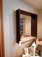 Front view of the square mirror mounted on a wall showing the deep wooden frame and reflection of a room with bookshelves and furniture.
