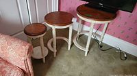 Set of 3 round wood nesting tables with white painted turned legs, natural wood finish tops, arranged from smallest to largest on carpeted floor near a TV and floral wallpapered wall.