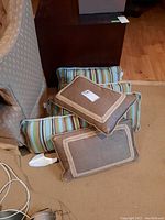 Six side pillows stacked on beige carpet with armchair in background, showing striped green-blue Sunbrella pillows and two brown pillows with cream trim.