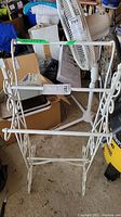 White metal quilt rack with two horizontal bars and decorative scrollwork on the sides, placed on a concrete floor surrounded by miscellaneous items including a fan and boxes.
