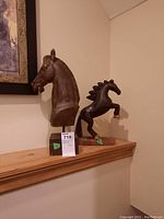 Two wood horse statues on a wooden shelf: a large horse head statue on a base and a smaller rearing horse statue on a base, against a wall with a framed picture.