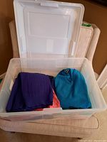 Open plastic storage bin filled with folded sweaters in various colors, placed on a beige armchair.