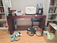 Photo showing a brown wood hallway table with a rectangular top and thick legs. Various household items like lamps, kitchen appliances (bread maker, coffee maker), and crockpots are placed on and around the table but are not included in the sale.