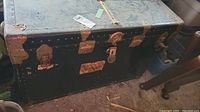 Front and top view of black vintage steamer trunk with metal latches and corner reinforcements. Shows surface wear, paint scratches, and chipping.