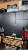 Front view of the black veneer wall unit showing grid of 20 square cubbies with doors and drawers, placed against wall in a cluttered storage setting.