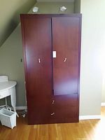 Frontal view of a tall wood wardrobe with two doors and two drawers at the bottom, placed against a wall. Shows minor scratches and white handles on doors and drawers.