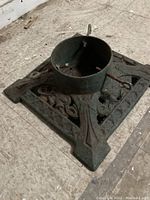 Angled view of a cast iron Christmas tree stand showing square base, central cylindrical tree holder, and securing screw rods with eyelet handles.