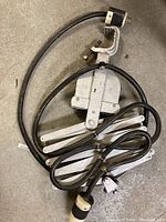 Photo of metal accordion clamp mechanism with attached black electrical cable and plug lying coiled on the ground.