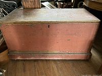 Front view of large antique wooden trunk painted red with dark trim, showing flat top and keyhole.