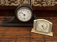 Two vintage clocks side by side on a wooden surface, one larger black mantel clock with round dial, one smaller beige tabletop clock with striped sides.