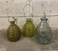 Three glass wasp catchers displayed side by side against a white brick wall, showing differences in shape and color.