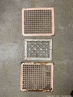 Top view of three metal floor grates: two pink square grates with grid patterns and one silver rectangular grate with geometric pattern.