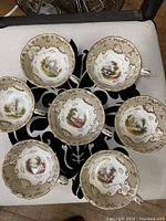 Overview photo of all seven vintage hand painted porcelain tea cups arranged with handles visible, showing distinct gold accents and scenic paintings inside each cup.