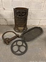 Antique cast iron stove part with decorative embossing, a cast iron tray, cast iron pulley-like device, and a round cast iron trivet arranged on the floor.