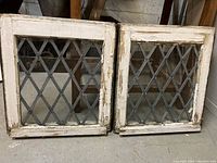 Pair of small square wooden framed leaded glass windows with diamond X pattern leaning against wall on floor. Paint on frames is chipped and worn.