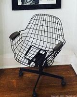 Frontal close-up of the black metal wire office chair showing grid wireframe seat, back, and brown padded armrests.