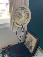 Photo showing decorative glass bowl with abstract multicolor pattern held by black metal stand next to two framed artworks on floor.