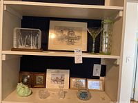 Two shelves showing assorted collectibles including framed artwork, glassware, and decor items.