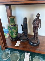 Photo showing all three items on a wooden shelf: green vase, tall wooden sculpture, bronze boy sculpture.