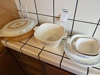 Three Corningware dishes and one lid on a tiled countertop. Dishes include a large round yellow-brown floral Spice of Life covered baking dish, a white square dish with Blue Cornflower pattern, and two smaller nested rectangular Blue Cornflower baking dishes.
