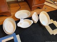9 pieces of white Corningware including 4 oval baking dishes, 4 round ramekins with spouts, and 1 oval casserole dish arranged on a rug on the floor.