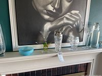 Crystal drinkware and decorative pieces displayed on a shelf under a black and white portrait painting. Includes glasses, goblets, vases, and a blue bowl.