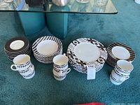 Photo of full dishware set on floor showing stacks of large plates, smaller plates, some bowls, and coffee mugs with black and white patterns.