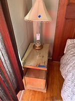 Wooden nightstand with wear marks and a brass lamp on top next to a bed.