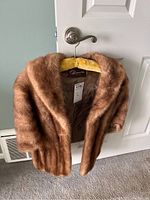 Front and angled view showing soft brown fur shawl with shawl collar hanging on yellow padded hanger against white door.