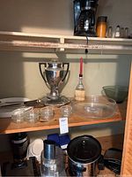 Wooden shelf with a silver trophy-style insulated pot, round and oval glass platters, four small round glass bowls, a food processor or juicer base, and a crockpot with glass lid below on lower shelf.