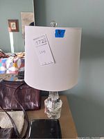 Front view of table lamp showing clear glass base, white lampshade, and lamp tag marked '1728'.